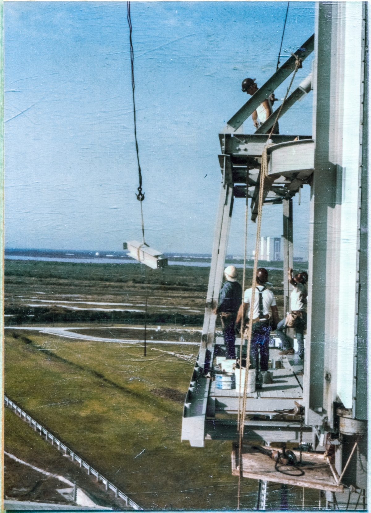Image 058. Union Ironworkers from Local 808 working for Ivey's Steel Erectors at Space Shuttle Launch Complex 39-B, Kennedy Space Center, Florida, prepare to receive an inbound steel beam, which will be attached to the reworked PBK & Contingency Platform which they are standing on, and above, out in front of the face of the Rotating Service Structure, an unimpeded 80 foot free drop above the concrete surface of the Pad Deck, beneath them. There is a story with the rework of this platforming, and it is not a pleasant one. Construction work, and Operations work too, in areas like this, is never to be taken for granted, and you must never let your guard down, lest you become an unwilling part of the “contingency” yourself. Photo by James MacLaren. Image 058. Union Ironworkers from Local 808 working for Ivey's Steel Erectors at Space Shuttle Launch Complex 39-B, Kennedy Space Center, Florida, prepare to receive an inbound steel beam, which will be attached to the reworked PBK & Contingency Platform which they are standing on, and above, out in front of the face of the Rotating Service Structure, an unimpeded 80 foot free drop above the concrete surface of the Pad Deck, beneath them. There is a story with the rework of this platforming, and it is not a pleasant one. Construction work, and Operations work too, in areas like this, is never to be taken for granted, and you must never let your guard down, lest you become an unwilling part of the “contingency” yourself. Photo by James MacLaren.